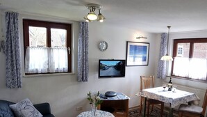 Ein Wohnzimmer mit einer Couch, einem Esstisch mit Stühlen, einem Fernseher, einer Uhr und einem Fenster mit Vorhängen.