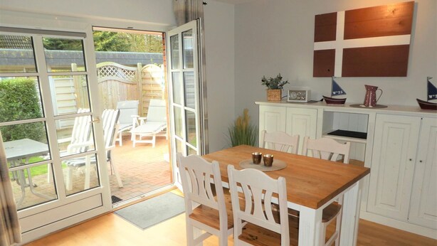 A dining area with a wooden table and white chairs, a kitchen with white cabinets, and a sliding glass door leading to an outdoor patio with chairs and a table. - Lüttje Fischerhuus - Holiday home Lüttje Fischerhuus: 400 m to the beach, terrace & (Wyk auf Föhr)