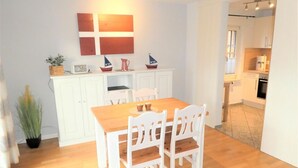 A dining area with a wooden table and white chairs, a kitchen in the background, and a wall-mounted artwork. - Lüttje Fischerhuus - Holiday home Lüttje Fischerhuus: 400 m to the beach, terrace & (Wyk auf Föhr)