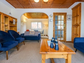 A cozy living room with a sofa, a wooden coffee table with binoculars, and a kitchen in the background. - Min Y Traeth, Bull Bay (Amlwch)