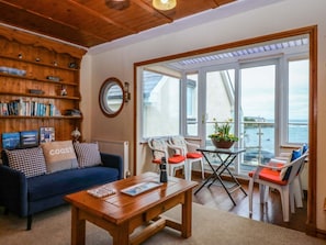 A cozy living room with a sofa, coffee table, and bookshelf, a balcony with chairs and a table, and a large window offering a view. - Min Y Traeth, Bull Bay (Amlwch)