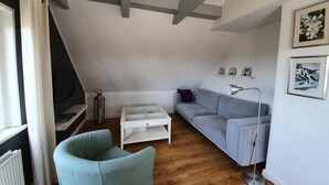 Un espacio tipo loft con techo inclinado, un sofá gris, un sillón azul claro, una mesa de centro blanca y una lámpara de pie.