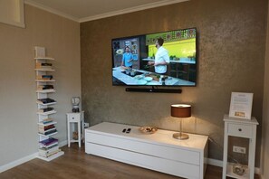 A living room with a flat-screen TV mounted on the wall, a white console table, a lamp, a bookshelf, and a small side table. - Austernperle - Holiday apartment Austernperle on Föhr - sauna, terrace & close to the beach (Wyk auf Föhr)