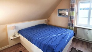 Un dormitori amb llit, una tauleta de nit, una finestra amb cortines, un radiador i un quadre a la paret.