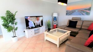 Ein Wohnzimmer mit Flachbildfernseher, einer Couch, einem Couchtisch, einem Blumentopf und einem Bild an der Wand.