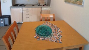 Eine kleine Küche mit Mikrowelle, Herd und Spüle. Ein Esstisch mit Stühlen und eine dekorative Schale sind vorhanden. Ein Gemälde hängt an der Wand.