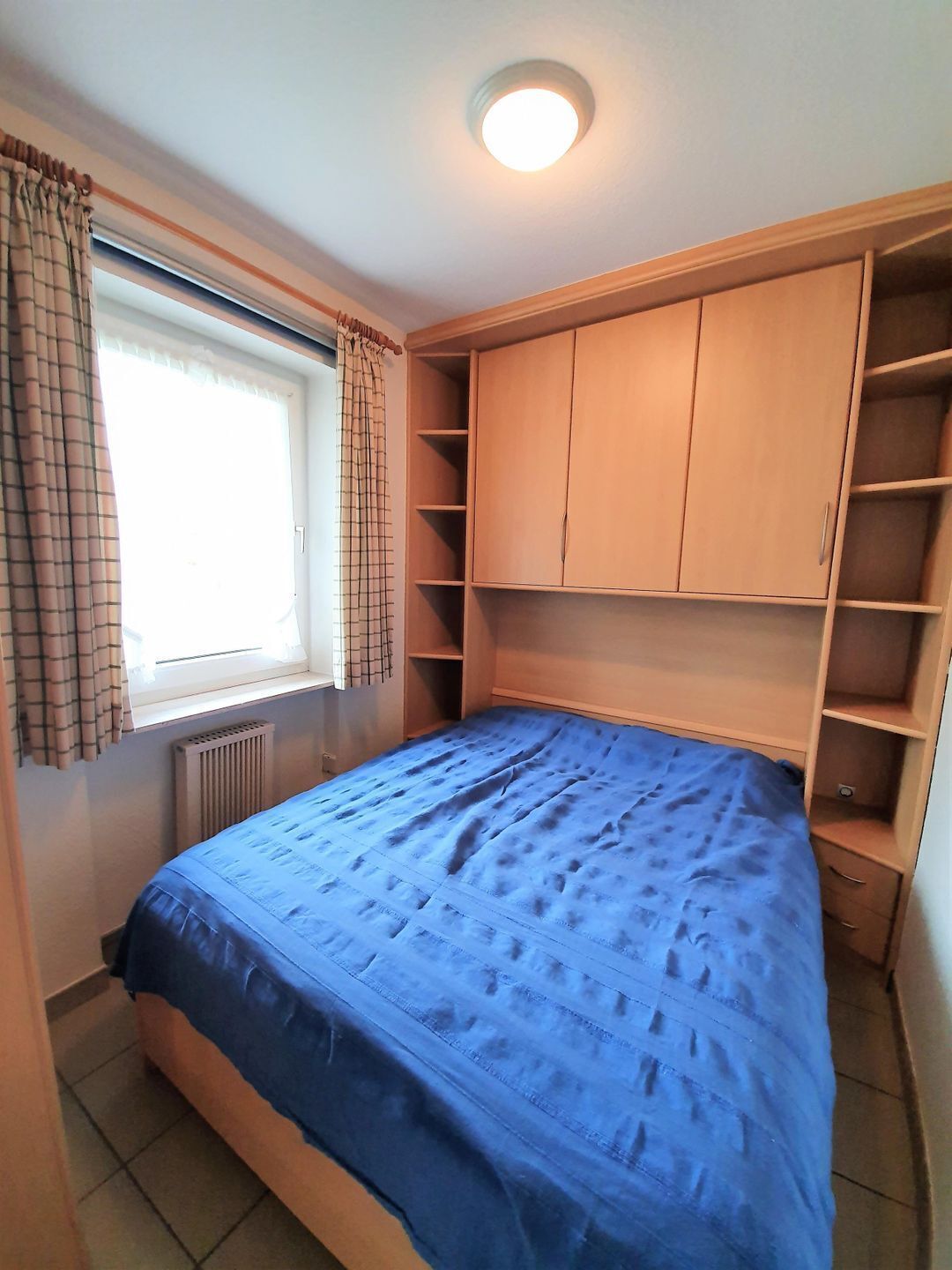 A bedroom with a bed, a window with curtains, a radiator, and built-in wooden shelving.