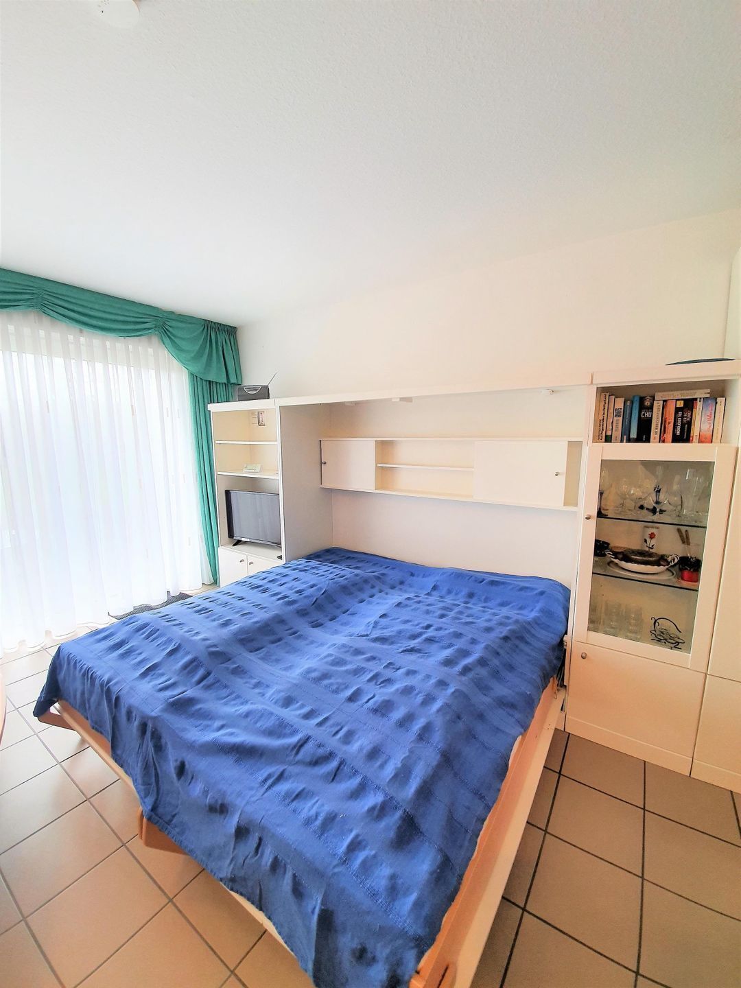 A bedroom with a bed, a desk, and a cabinet with books and decorative items.