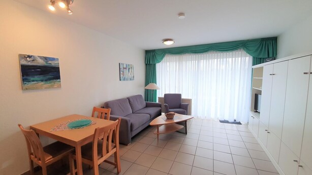 Ein Wohnzimmer mit einer Couch, einem Esstisch mit Stühlen, einer Lampe und Blick ins Freie durch ein Fenster mit Vorhängen.
