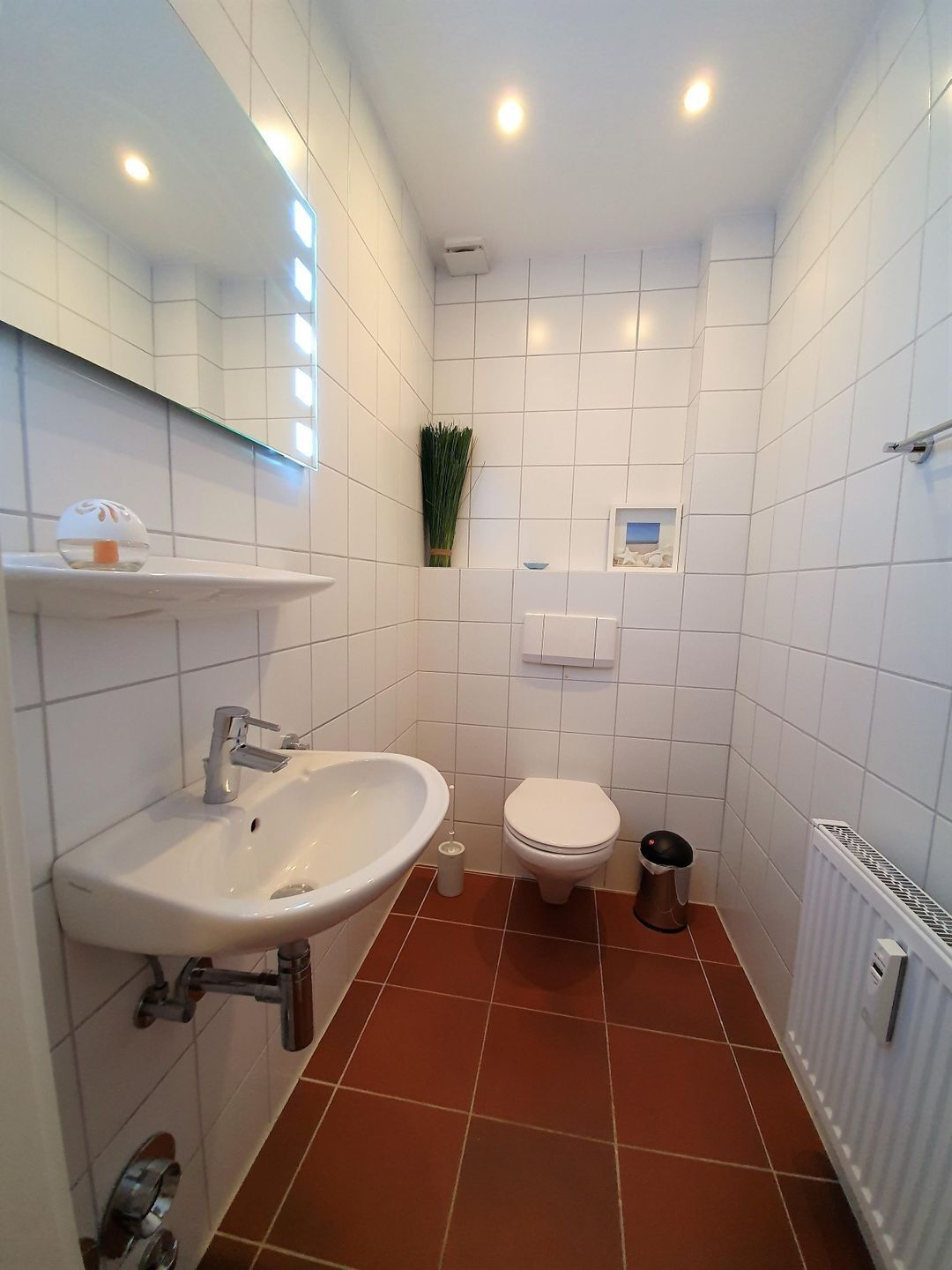 A bathroom with white tiles, a sink, a toilet, a radiator, and a small shelf with a decorative item.