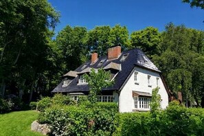 Una casa de dues plantes amb teulada a dos aiguavessos, envoltada de vegetació exuberant.