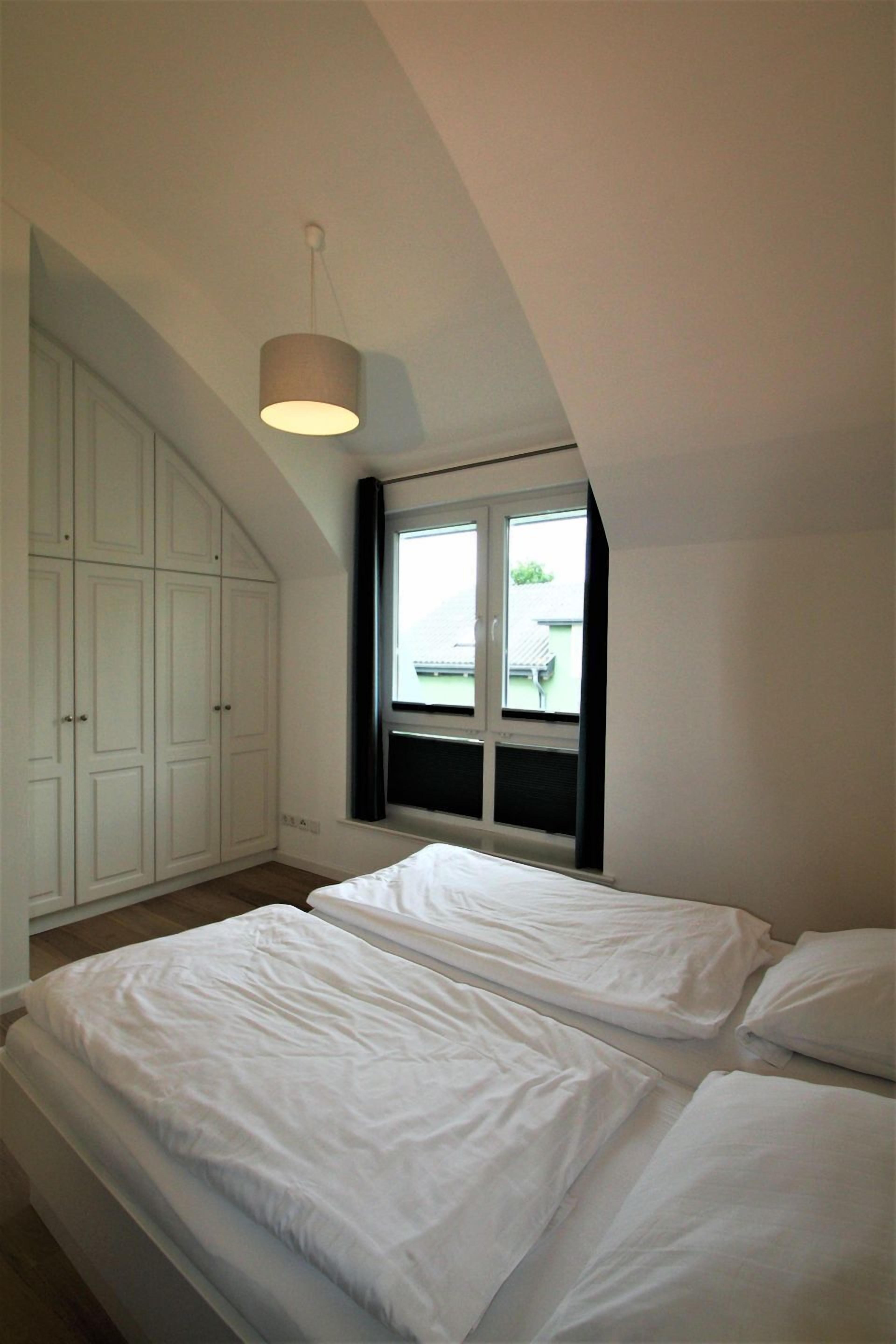 Ein Schlafzimmer mit schrägen Wänden, einem Fenster mit Jalousien, einem Bett mit weißer Bettwäsche und einer Hängeleuchte.