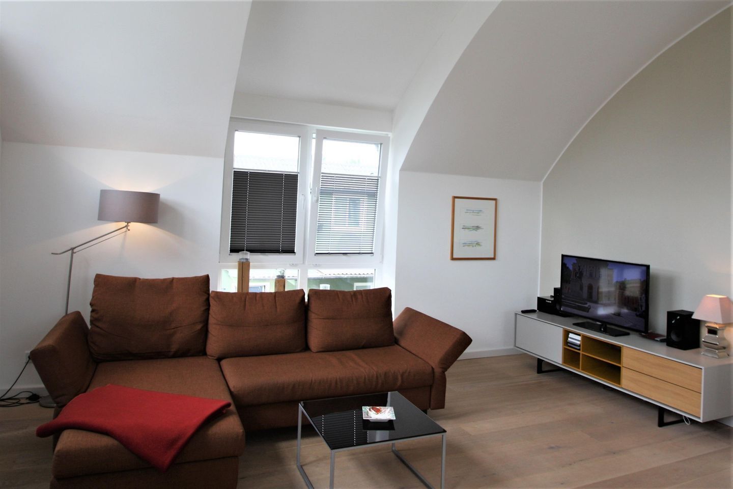 Un salon moderne avec un canapé marron, une table basse en verre, un téléviseur posé sur un meuble, une lampe sur pied et une fenêtre avec des stores.