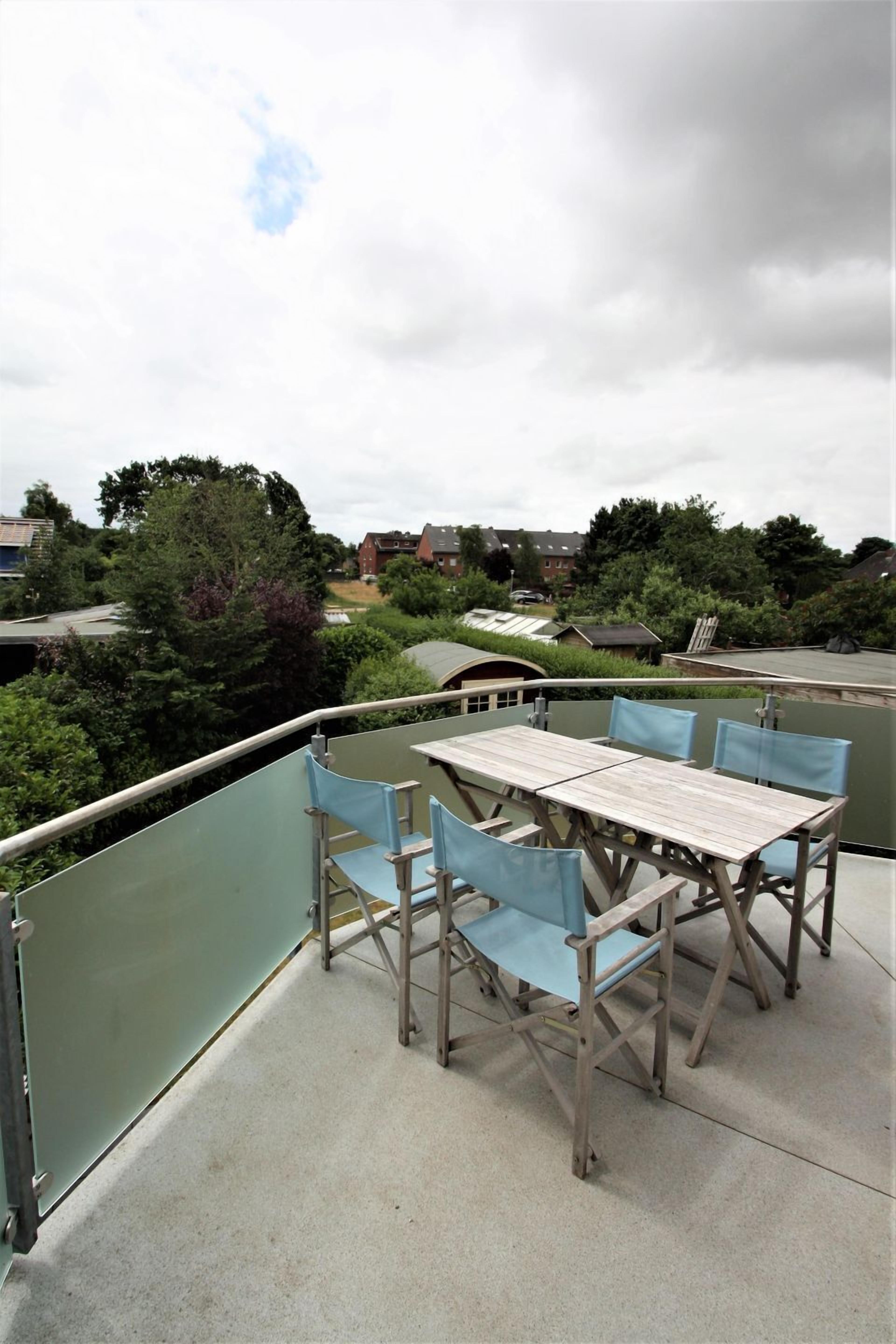 Een balkon met een tafel, stoelen, een glazen hekwerk en uitzicht op groen en gebouwen.