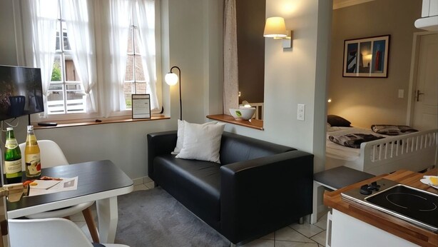 Een knus woongedeelte met een bank, een eettafel, een televisie, een kitchenette en een raam met gordijnen.