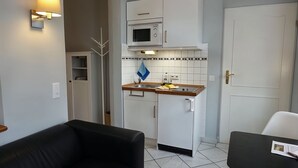 Een kleine keuken met een magnetron, fornuis en witte kastjes. Er staat een zwarte bank en een klein tafeltje met een menukaart.