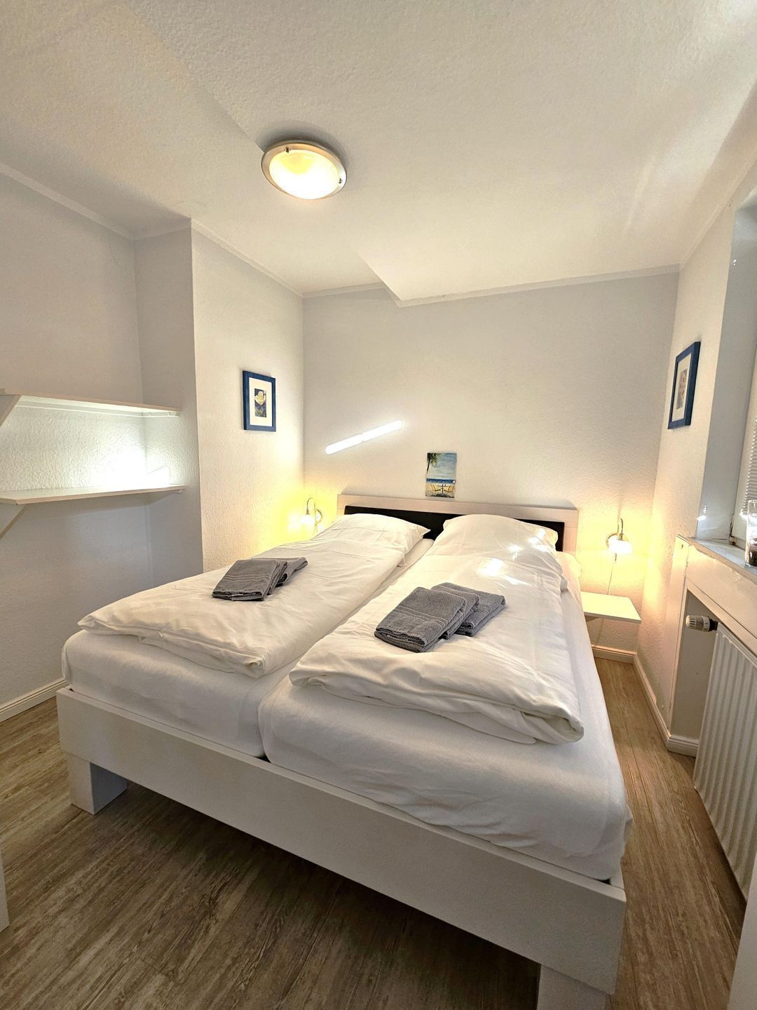 Un lit double avec du linge de lit blanc, deux lampes de chevet, un luminaire au plafond et un tableau encadré au mur.