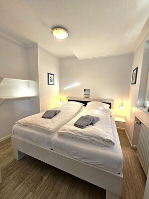 A double bed with white linens, two bedside lamps, a ceiling light, and a framed picture on the wall. - Kiebitz - Allergy-friendly vacation apartment Kiebitz with balcony near the beach in (Wyk auf Föhr)