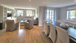 A spacious living area with a sofa, chairs, a coffee table, and a dining table with chairs. There is a television mounted on a wall, and a window with curtains. - Golfoase 1 - Semi-detached house for 6 people - Golfoase 1: Semi-detached house for 6 people (Oevenum)