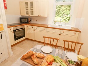 A kitchen with a microwave, oven, and wooden cabinets. There is a dining table with plates, glasses, and a variety of food items. - Seaview Retreat in Coverack (Coverack)