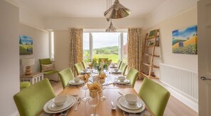 A dining room with a table set for six, green chairs, a chandelier, and a view of the countryside. - Hazelseat House, Ulverston (Ulverston)