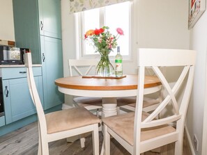 A kitchen with teal cabinets, a white dining table with two chairs, and a window with flowers. - Hunrosa: Charming Home in St Day (Redruth)