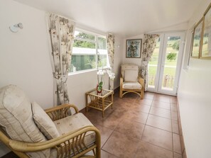 A room with a wicker chair, a cushioned chair, a small table with a plant, and a view through a glass door to the outside. - The Farmhouse, Ledsham (Ledbury)
