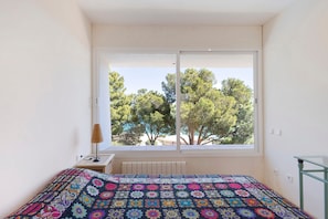 A bedroom with a large window, a bed with a colorful quilt, a bedside table with a lamp, and a radiator.