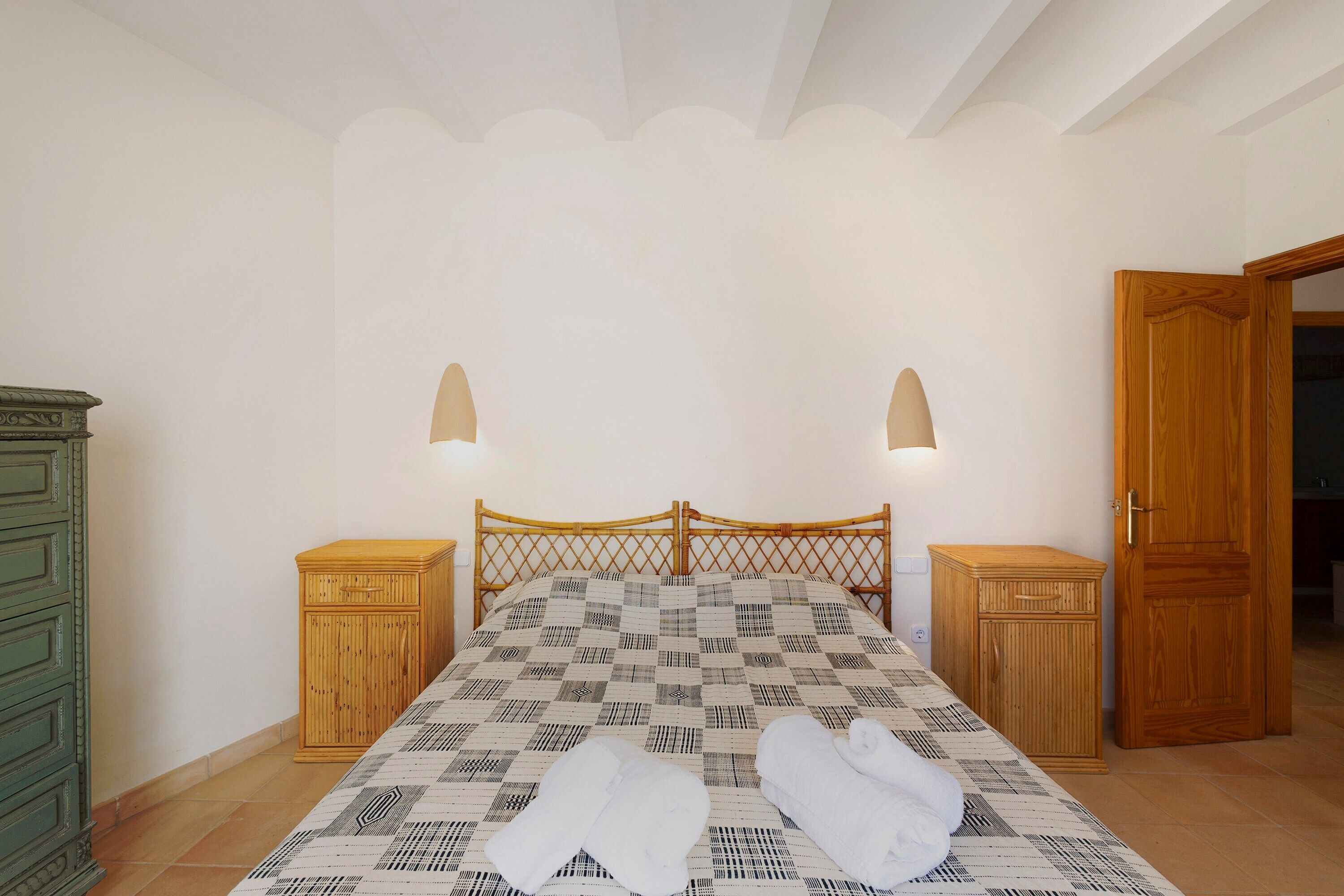 A bedroom with a checkered bedspread, two bedside tables, a wooden wardrobe, and a door leading to another room.