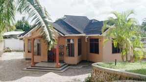 A single-story house with a tiled roof, a porch with a palm tree, and a stone wall.
