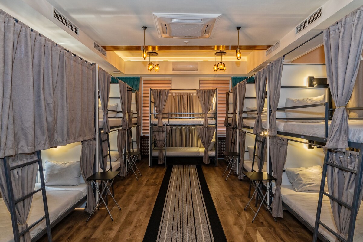 A dormitory room with bunk beds, a central seating area, and a wooden floor.