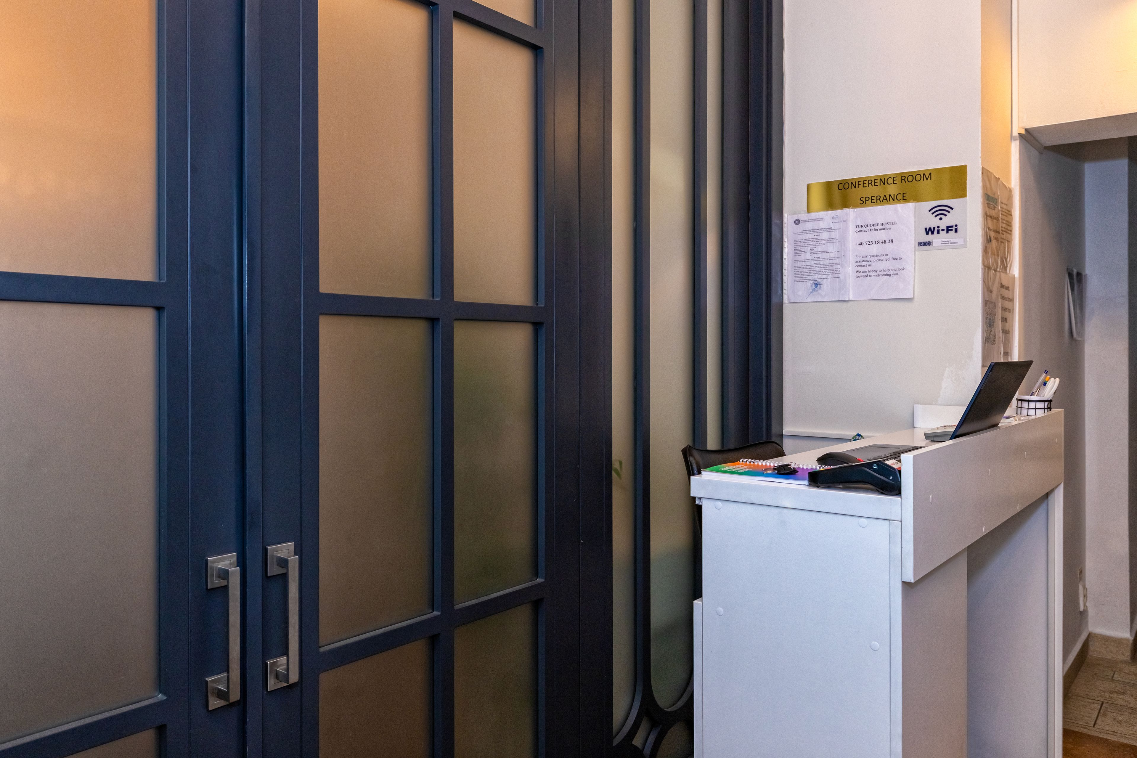 Ein Empfangsschalter mit einem Laptop, einem Telefon und einem Notizblock. Ein Schild weist auf einen Konferenzraum und kostenloses WLAN hin.