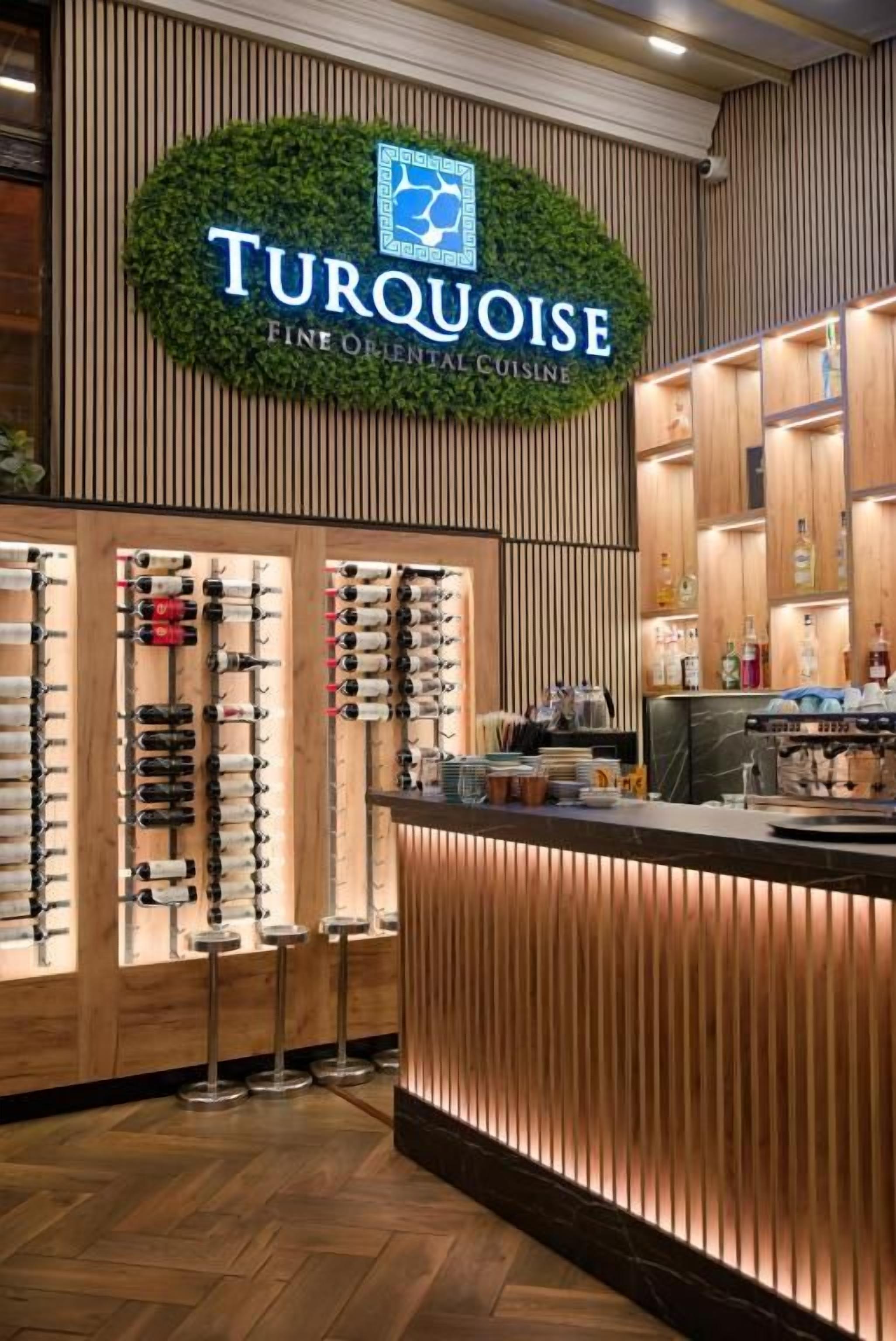 L’intérieur d’un restaurant, avec une enseigne indiquant « Cuisine orientale raffinée Turquoise », des lambris en bois et un présentoir à vins.