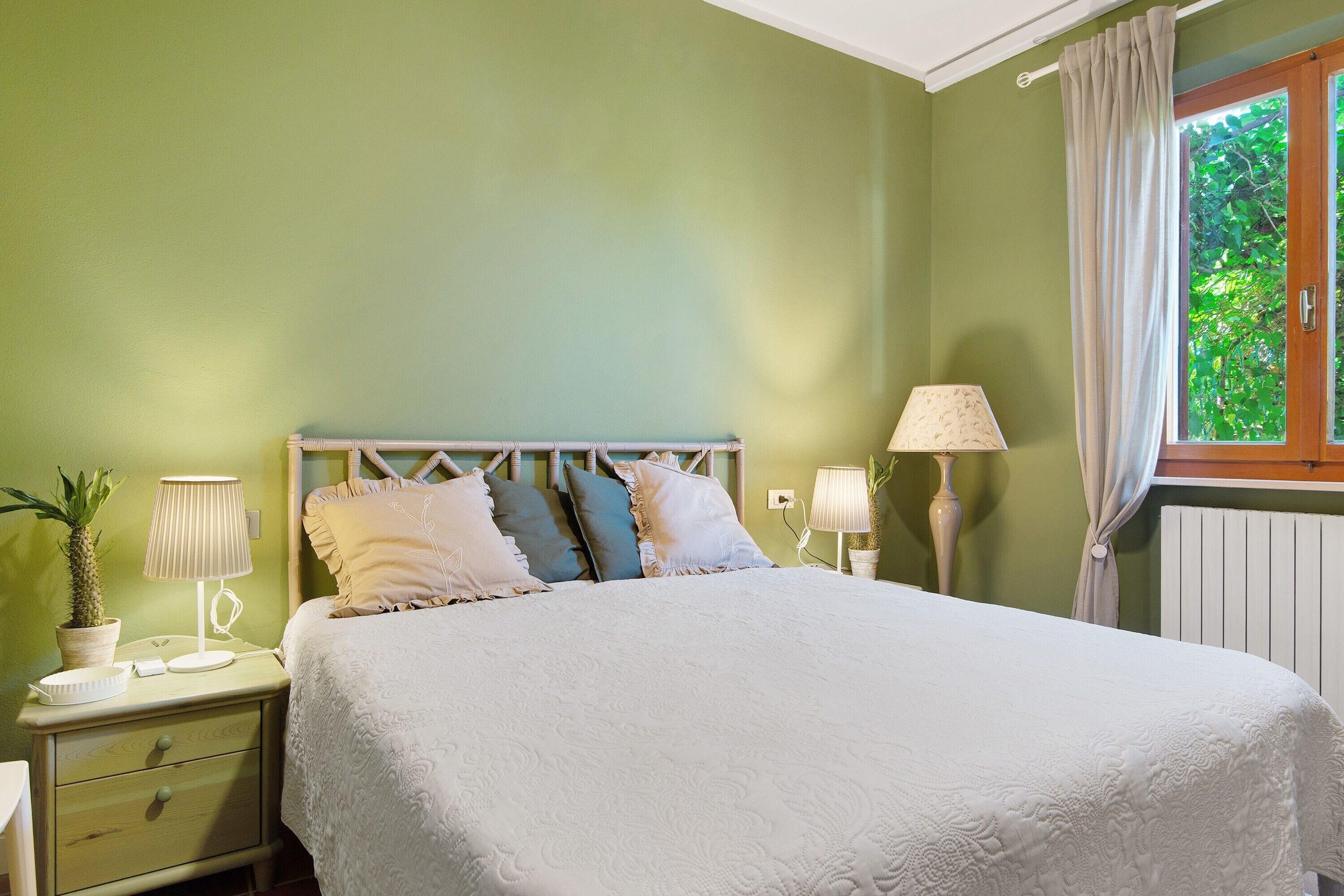Une chambre à coucher avec un lit, deux tables de chevet, une fenêtre avec des rideaux et une plante en pot.