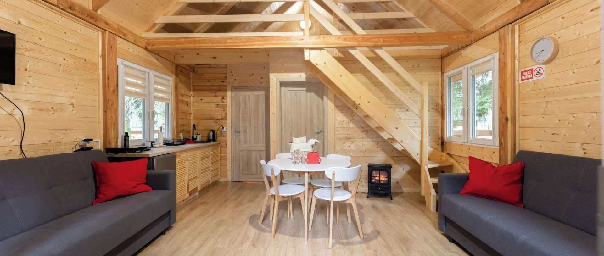 A wooden cabin interior with a dining area, two sofas, a staircase, and a fireplace.