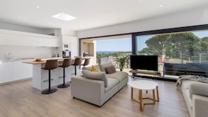 A modern living space with a kitchen, bar area, seating, and a large window with a view. - Villa con piscina, aire acondicionado y vistas al mar (Pineda Pals)