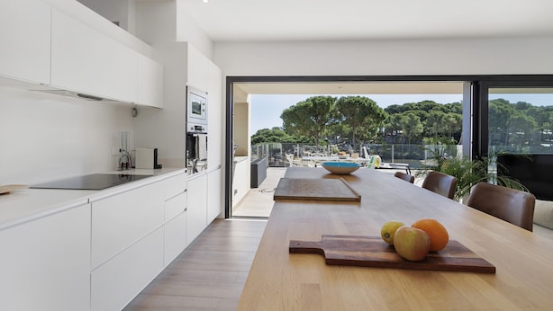A modern kitchen with white cabinetry, a wooden dining table with fruits, and a view of trees through large windows. - Villa con piscina, aire acondicionado y vistas al mar (Pineda Pals)