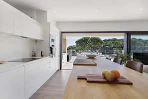 A modern kitchen with white cabinetry, a wooden dining table with fruits, and a view of trees through large windows.