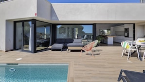 A modern house with a pool, outdoor seating, and a kitchen visible through glass doors. - Villa con piscina, aire acondicionado y vistas al mar (Pineda Pals)