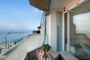 Un balcón con vista al mar, una mesa con frutas y una botella, y un sofá.