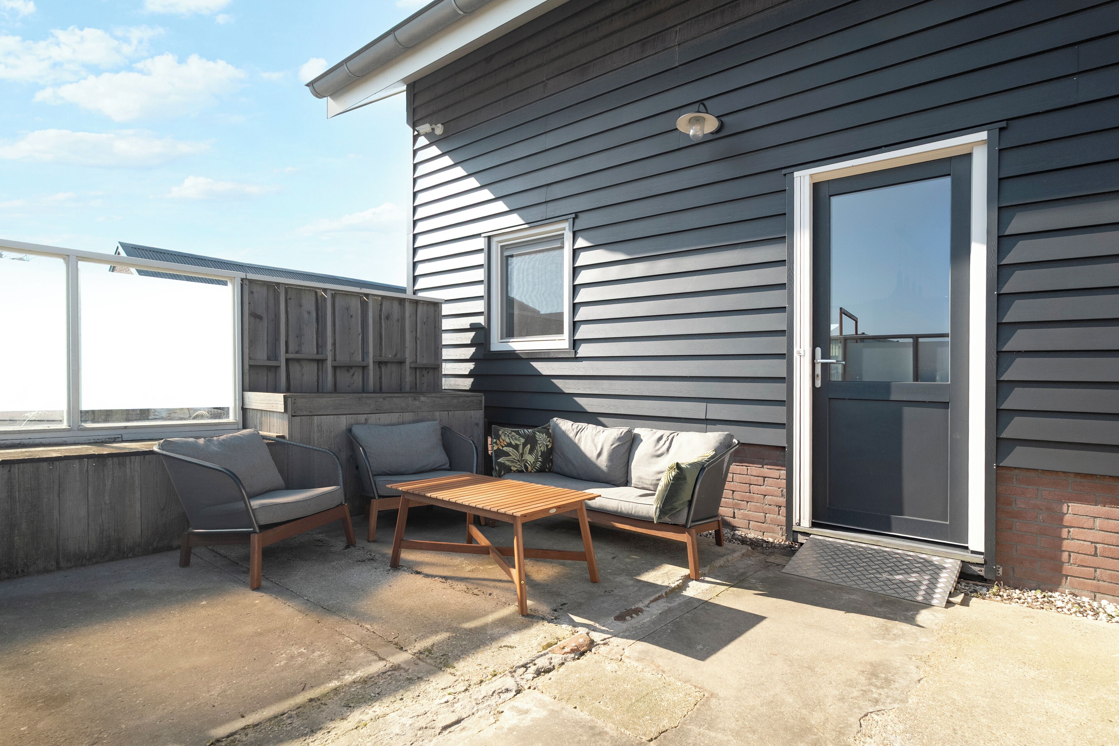 A patio with a wooden table and two grey sofas, a sliding glass door, and a small window.