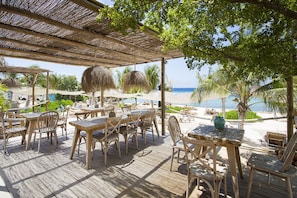 A wooden deck with tables and chairs, overlooking a beach with palm trees and a clear view of the ocean. - peaceful Garden Villa - Private Pool (Jan Thiel)