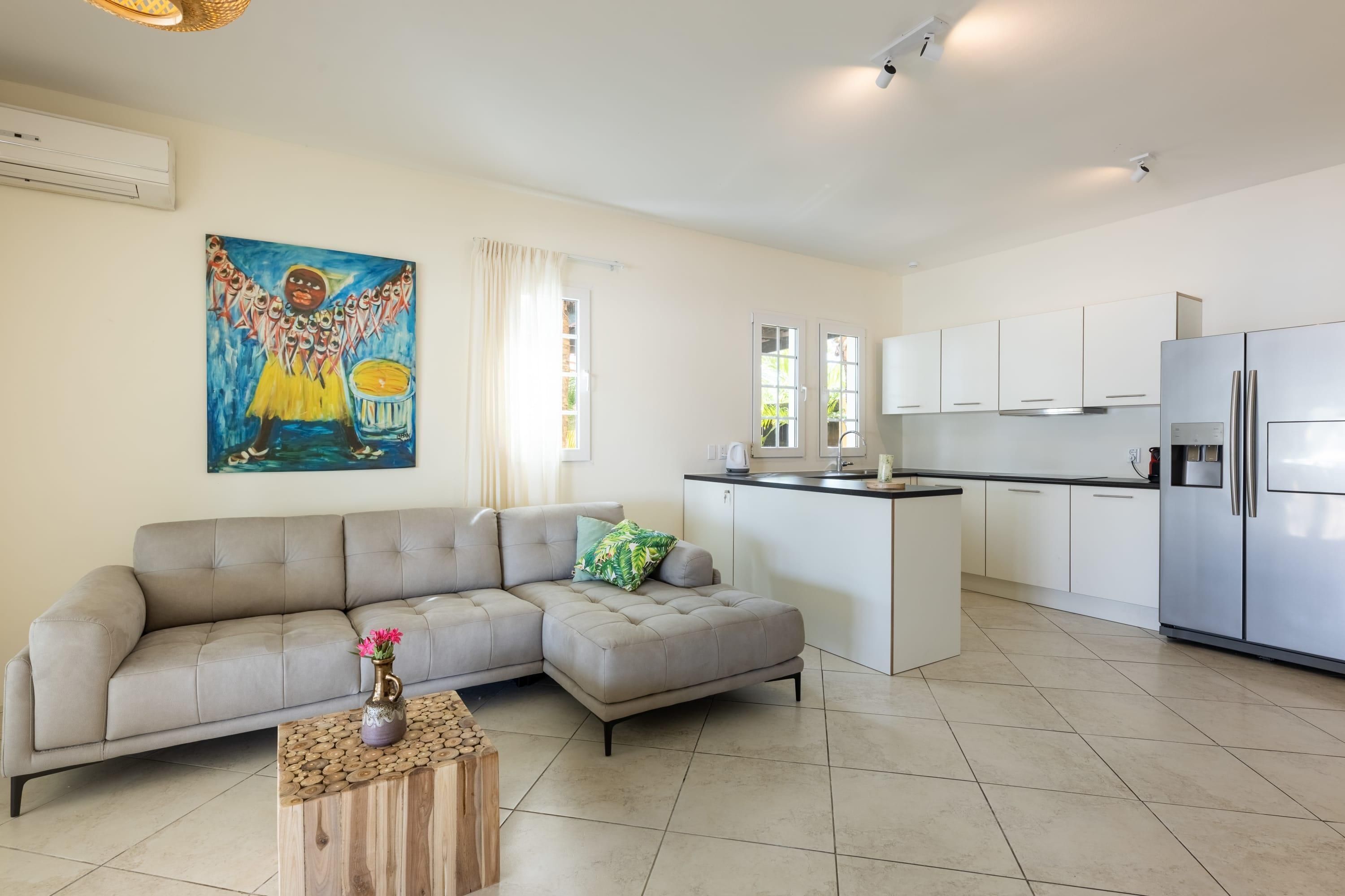 A modern living room with a sofa, a coffee table, a kitchen area with white cabinets and a refrigerator, and a painting on the wall.