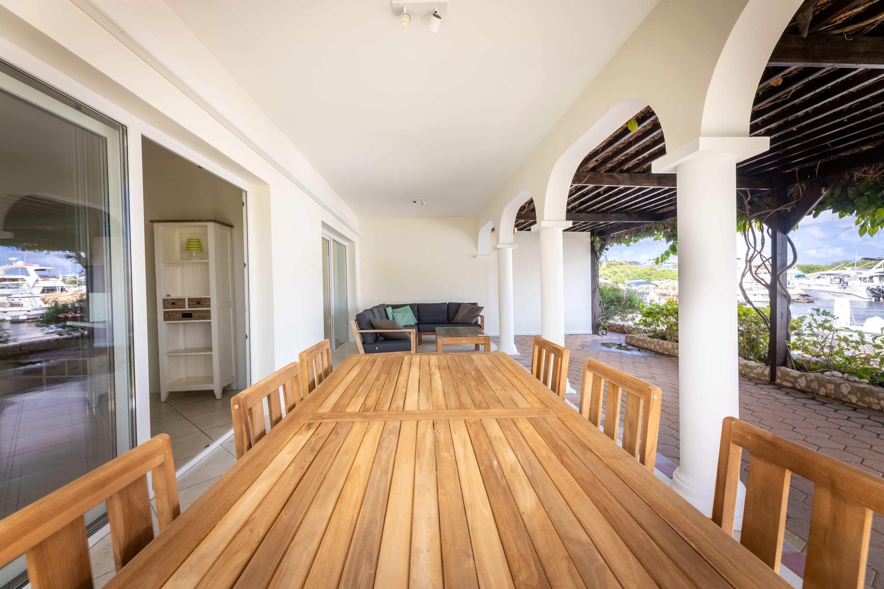 Una mesa de madera con sillas, un sofá y un ventanal de vidrio que da a un balcón con vistas a barcos.