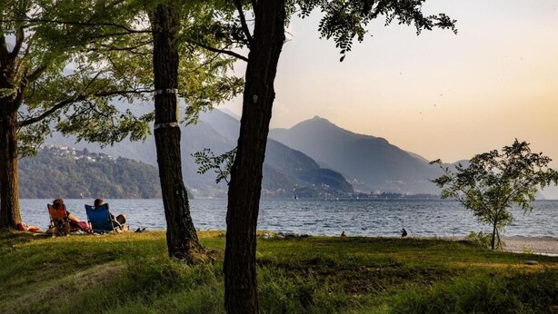 Un paysage au bord d’un lac, avec des arbres, une personne assise sur une chaise et une chaîne de montagnes en arrière-plan.