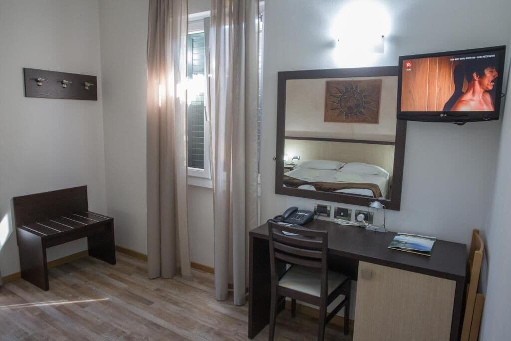 A hotel room with a bed, a desk with a chair, a television mounted above the bed, and a window with curtains.