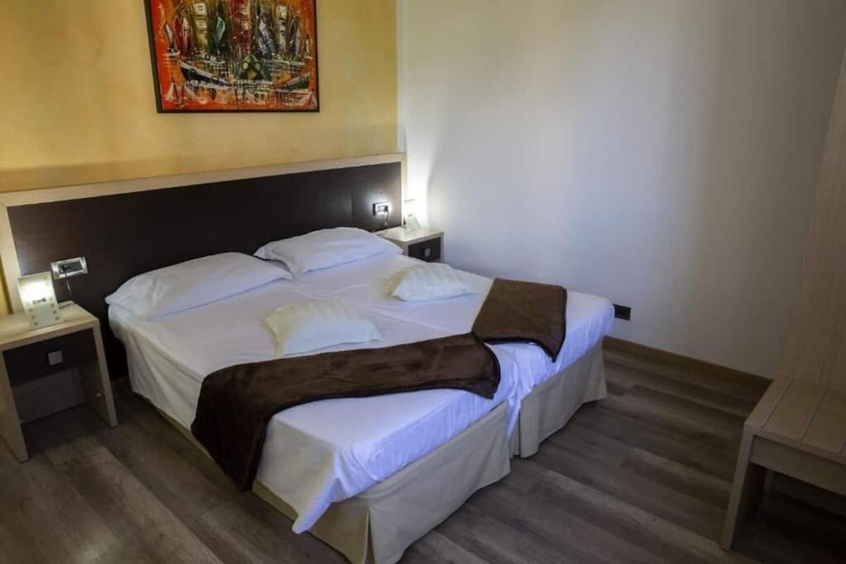 Un cuarto de hotel con una cama grande, dos mesitas de noche, un buró, un cuadro en la pared y piso de madera.