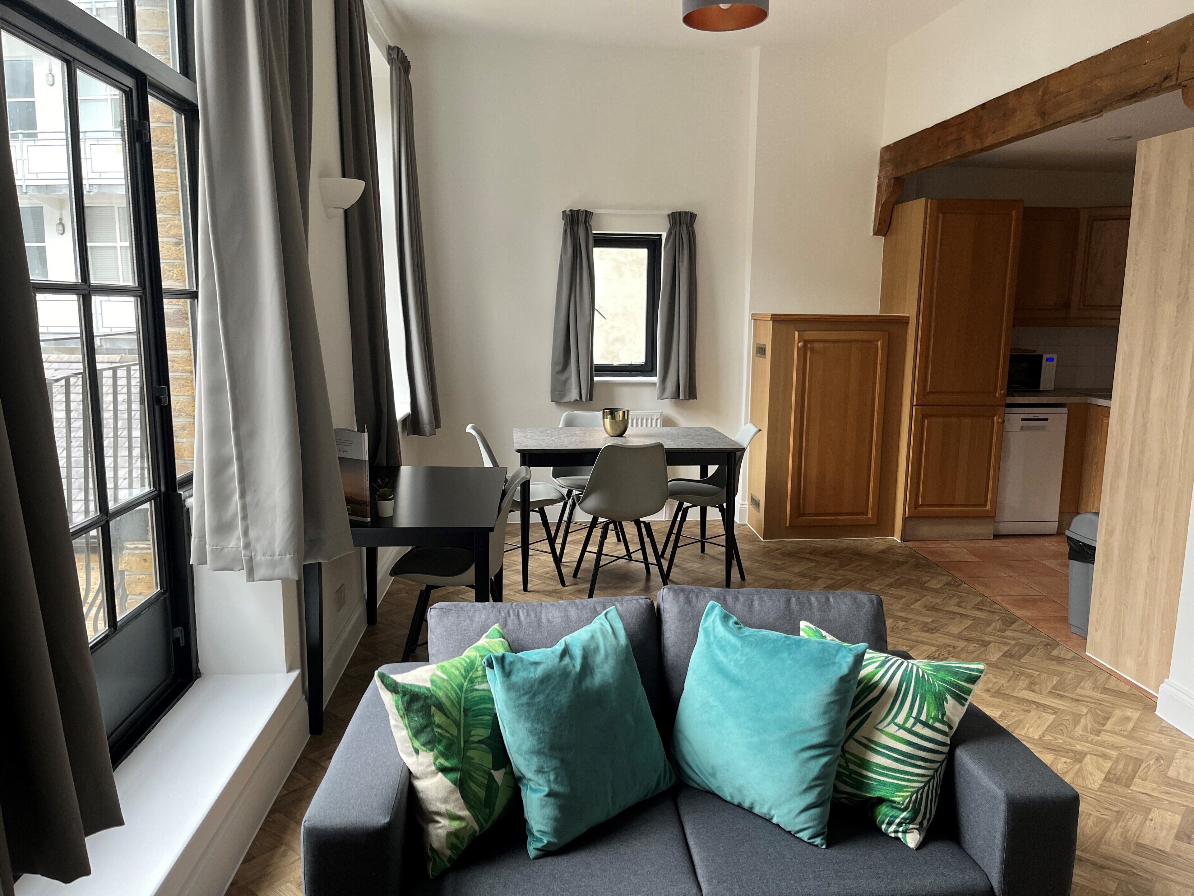 En moderne stue med sofa, spisebord med stole, et køkkenområde og store vinduer med gardiner.