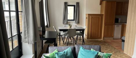 En moderne stue med sofa, spisebord med stole, et køkkenområde og store vinduer med gardiner.