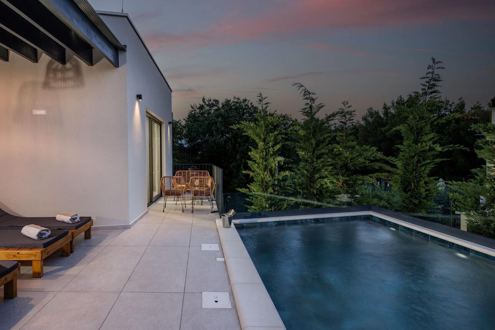 A modern outdoor pool area with lounge chairs and a table, surrounded by trees.